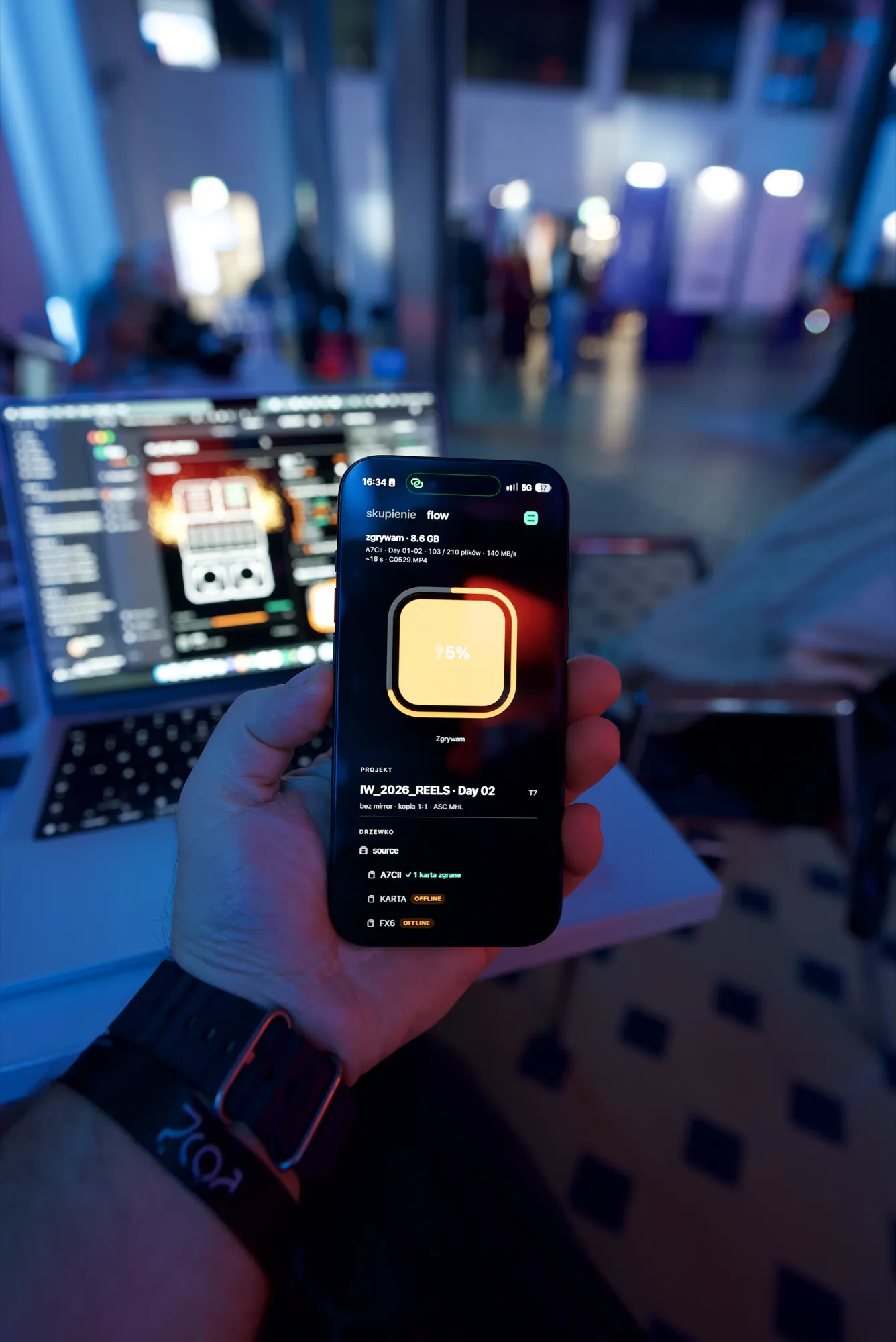 iPhone in hand shows an A7C II card being ingested, MacBook and the event hall in the background